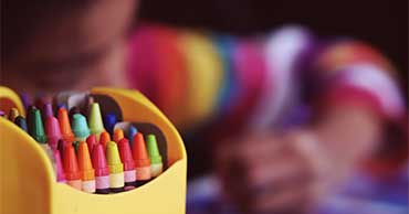 En la vanguardia, una caja de crayones, en el fondo un niño coloreado.