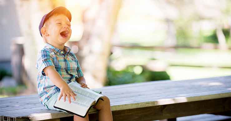 Child laughing heartily while sitting on a bench reading a book outside.