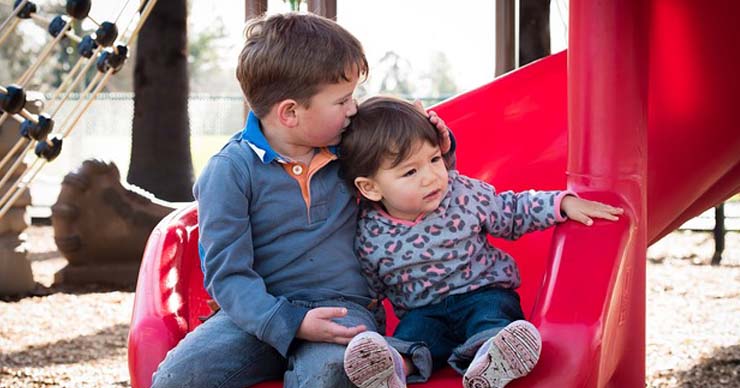 A older boy is giving a younger girl a hug at the bottom of a slide.