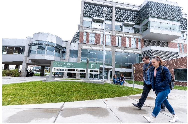 Two UVU students walk in front of the Fulton Library, with a library sign and two chatting students in the background.  