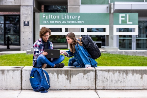 Two UVU students sit in front of an entrance to the Fulton Library. One student  holds a laptop, while the other points at the screen