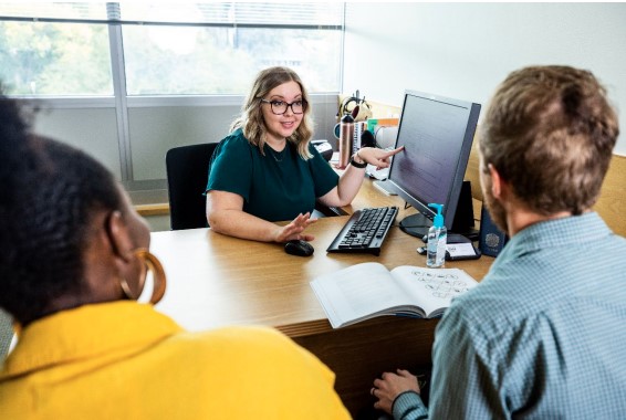A librarian points at a computer screen and provides research help to two students seated across a desk.