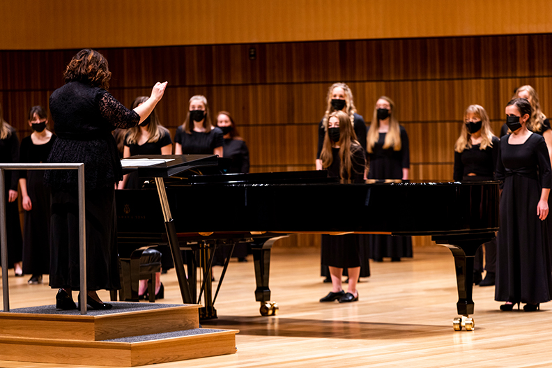 A woman conducts the women's choir, all dressed in black dresses with black masks