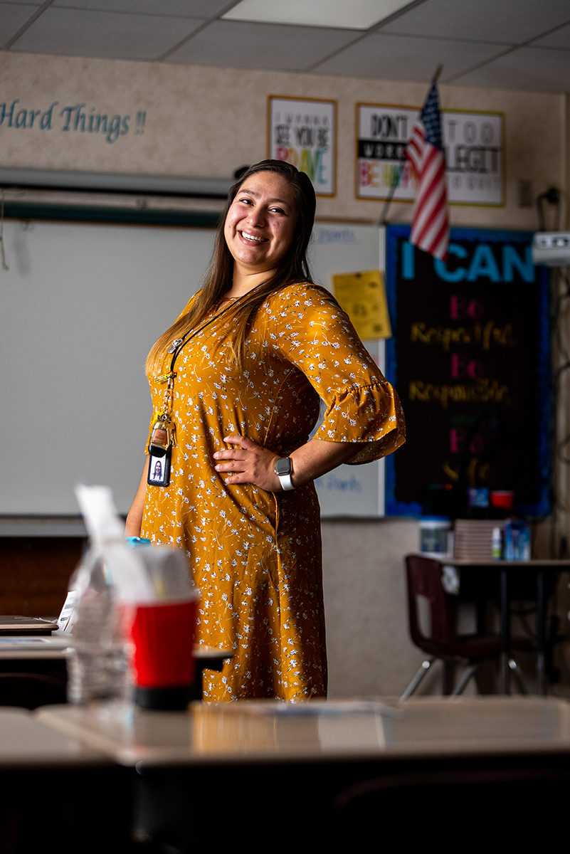 Crystal Sedano smiles with her hand on her hip in her classroom wearing a yellow dress with flowers on it