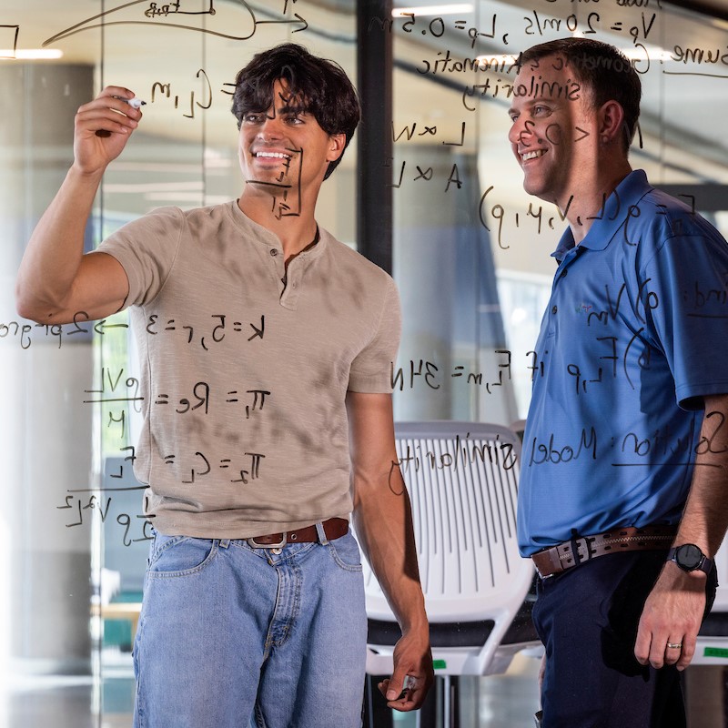 student writing on a board while a professor checks their work