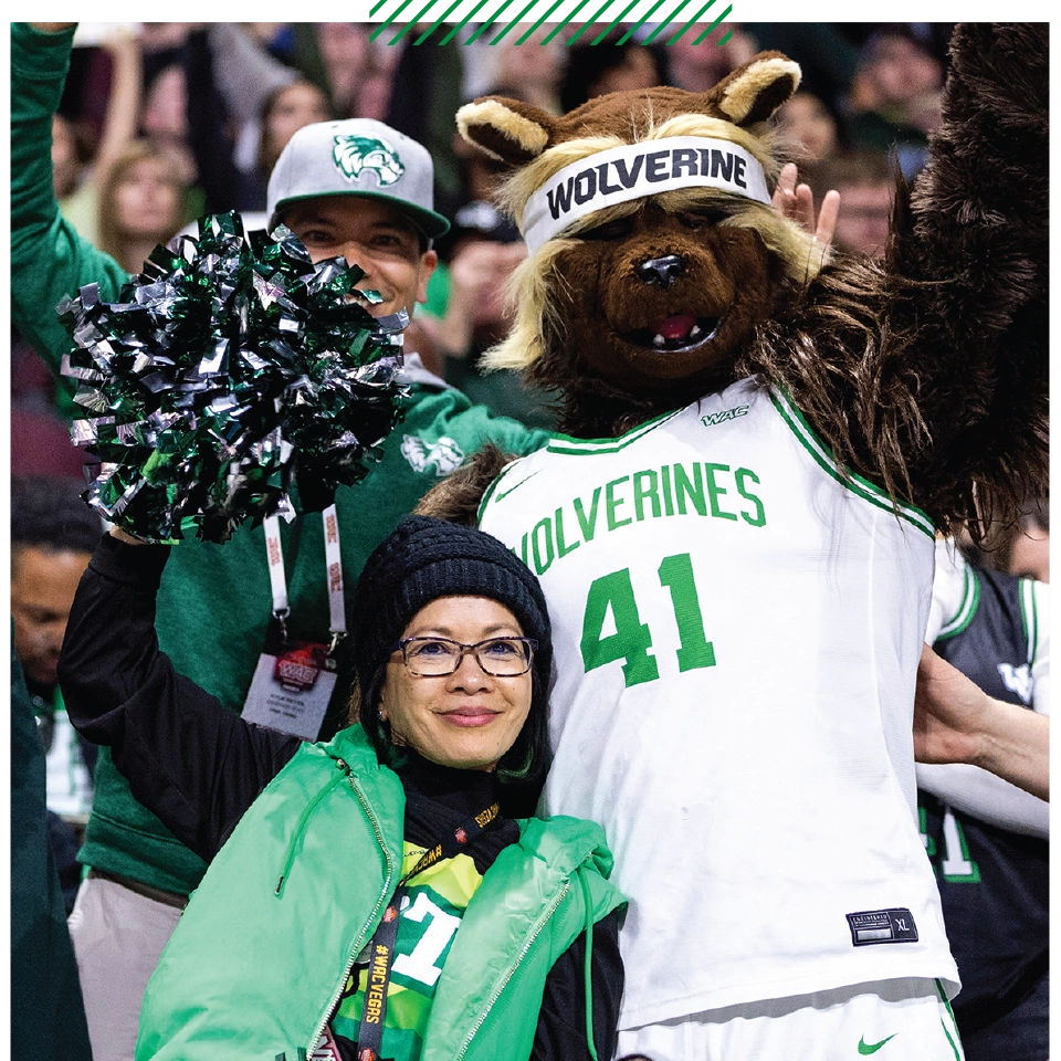 UVU President Astrid S. Tuminez, wearing a black beanie and green vest and holding pom-poms, poses with Willy the Wolverine in front of a crowd at a UVU sports event. 