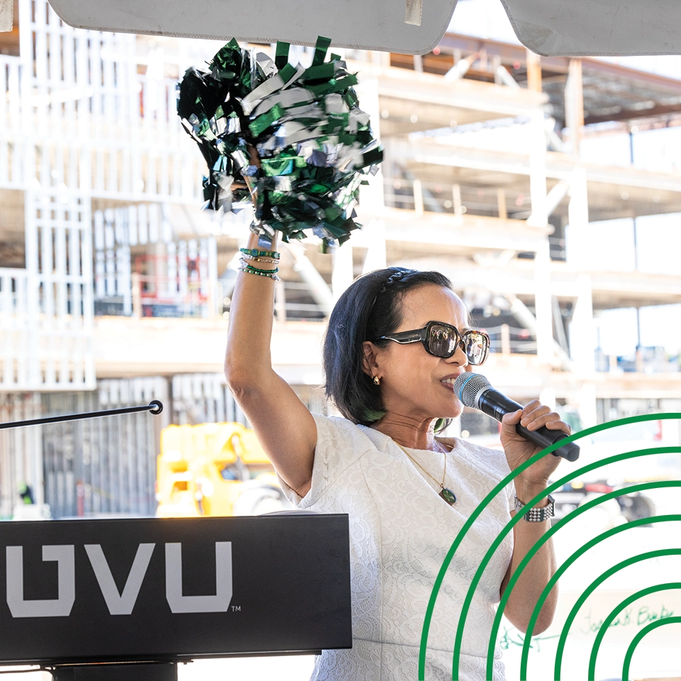 President Tuminez, wearing sunglasses and a white blouse, holds a pom-pom and speaks into a microphone at an event celebrating the construction of a campus building. 