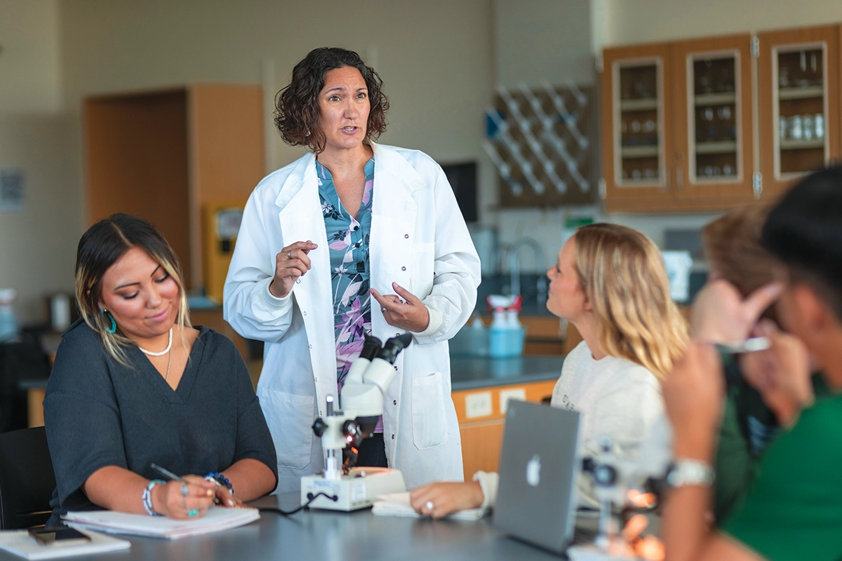 A UVU professor wearing a white lab coat teaches a class as a student takes notes. A UVU professor wearing a white lab coat teaches a class as a student takes notes.