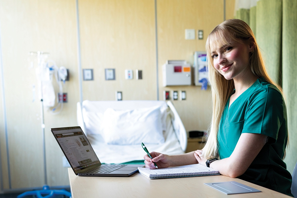A UVU nursing student wearing green scrubs sits at a desk and writes in a notebook. A UVU nursing student wearing green scrubs sits at a desk and writes in a notebook.