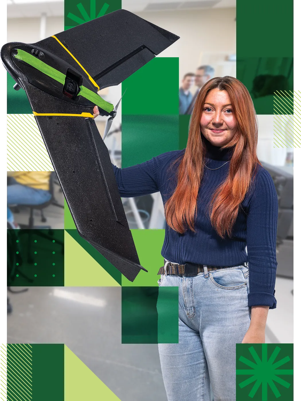 A UVU student with long red hair, wearing a navy turtleneck sweater and light blue jeans, holds up a black flying machine