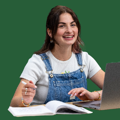 A UVU student in denim overalls and a white T-shirt smiles while taking notes in a lecture
