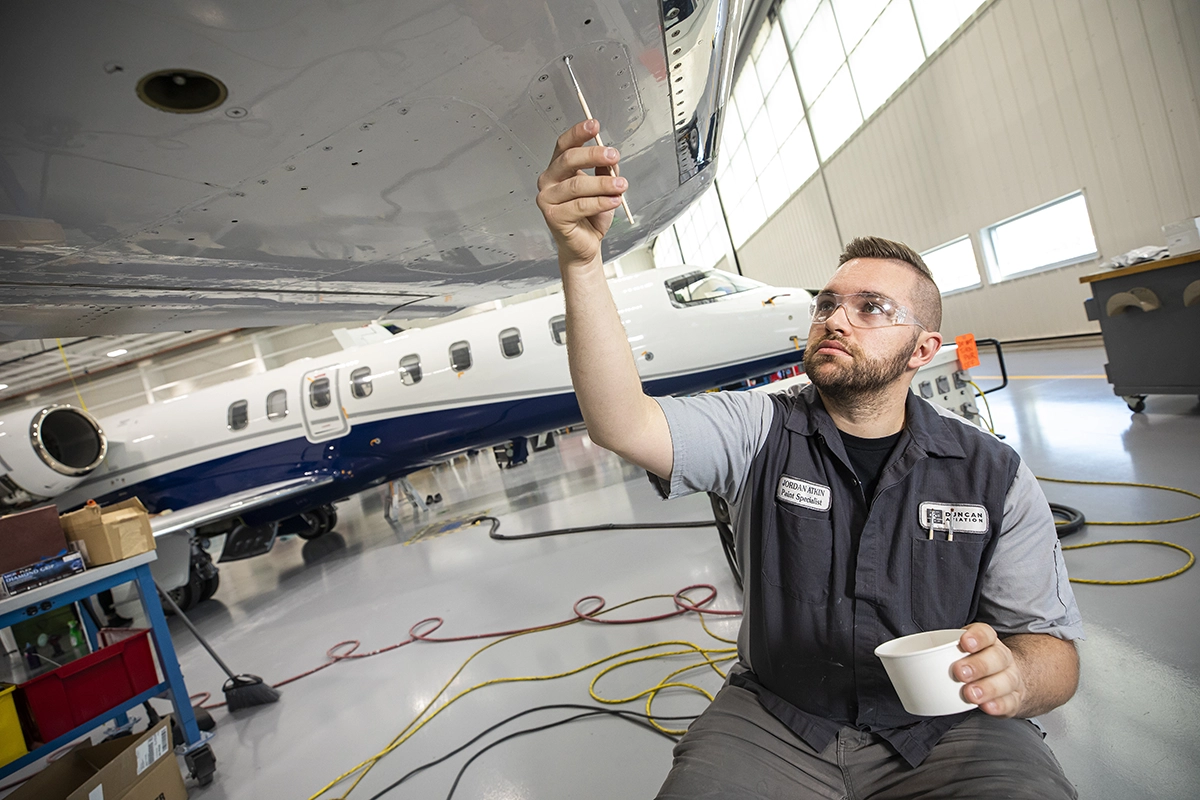 A man in a Duncan Aviation vest and safety goggles applies paint to an aircraft. A man in a Duncan Aviation vest and safety goggles applies paint to an aircraft.