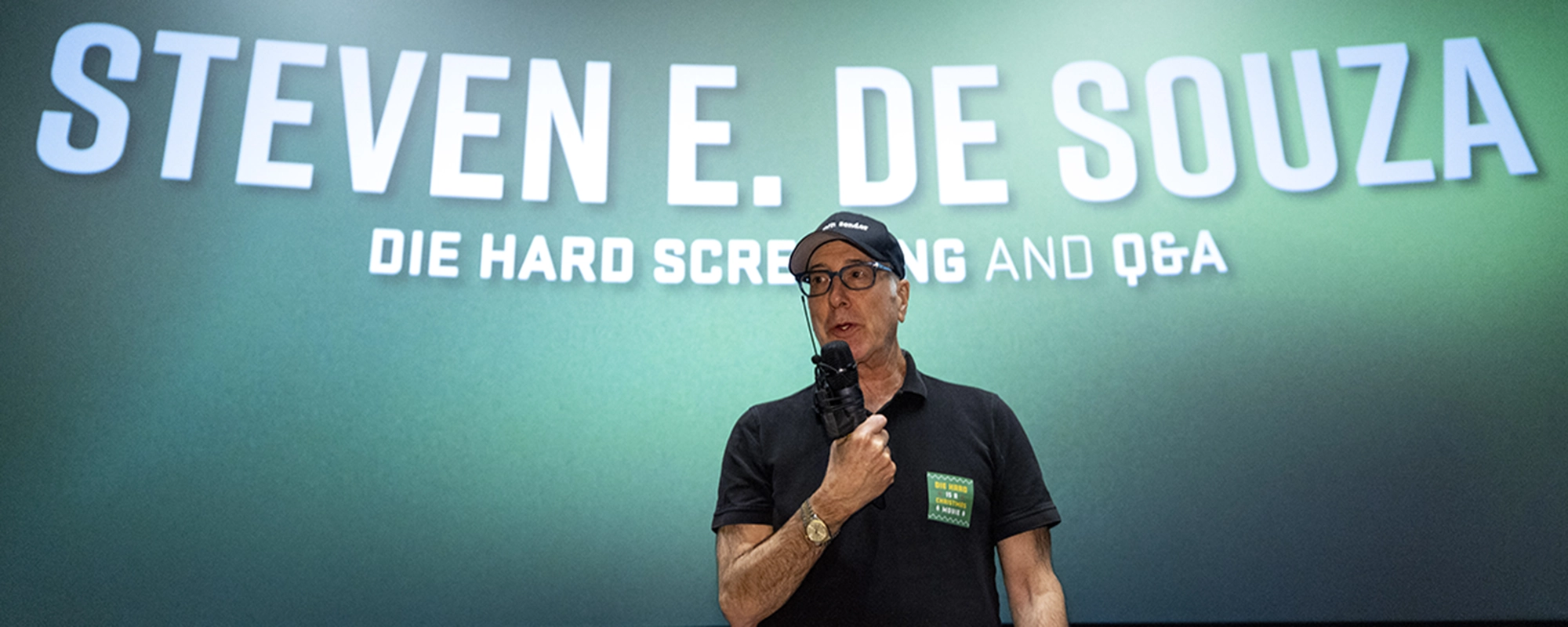 Screenwriter Steven E. de Souza, wearing a black polo shirt, black cap, and black glasses, holds a microphone while speaking to an audience. Screenwriter Steven E. de Souza, wearing a black polo shirt, black cap, and black glasses, holds a microphone while speaking to an audience.