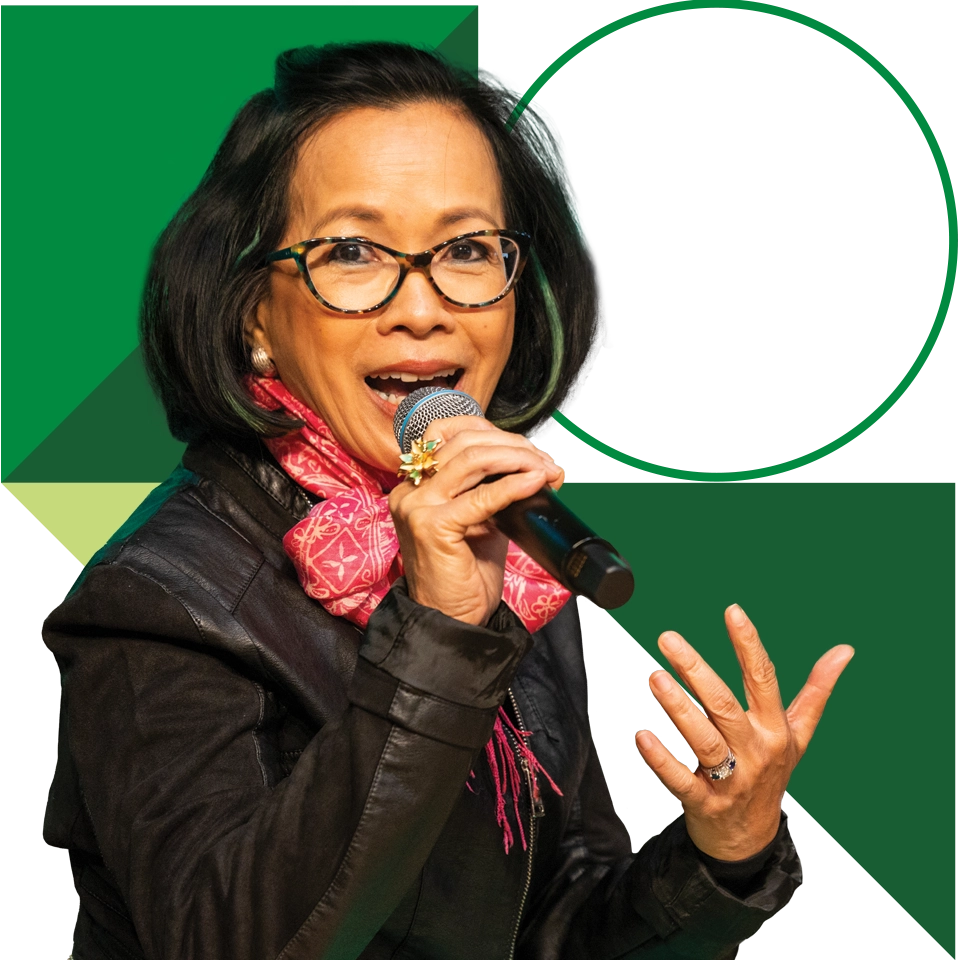 President Tuminez, wearing a black leather jacket and a red scarf, speaks into a microphone. President Tuminez, wearing a black leather jacket and a red scarf, speaks into a microphone.