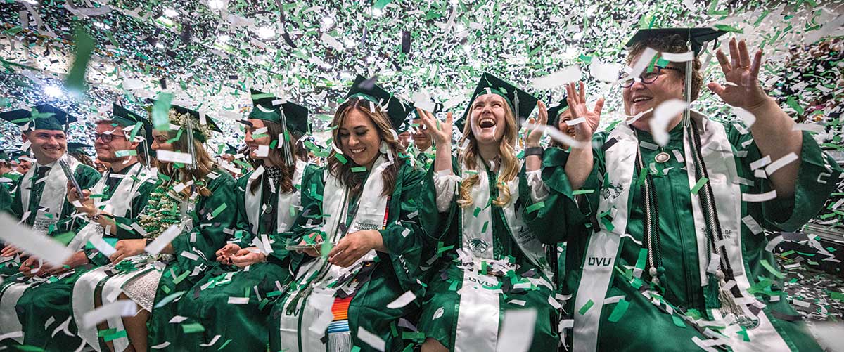 A UVU student, who is wearing a green cap and green graduation regalia, smiles while sitting in a crowd of other graduates at UVU’s 2023 commencement ceremony. 
