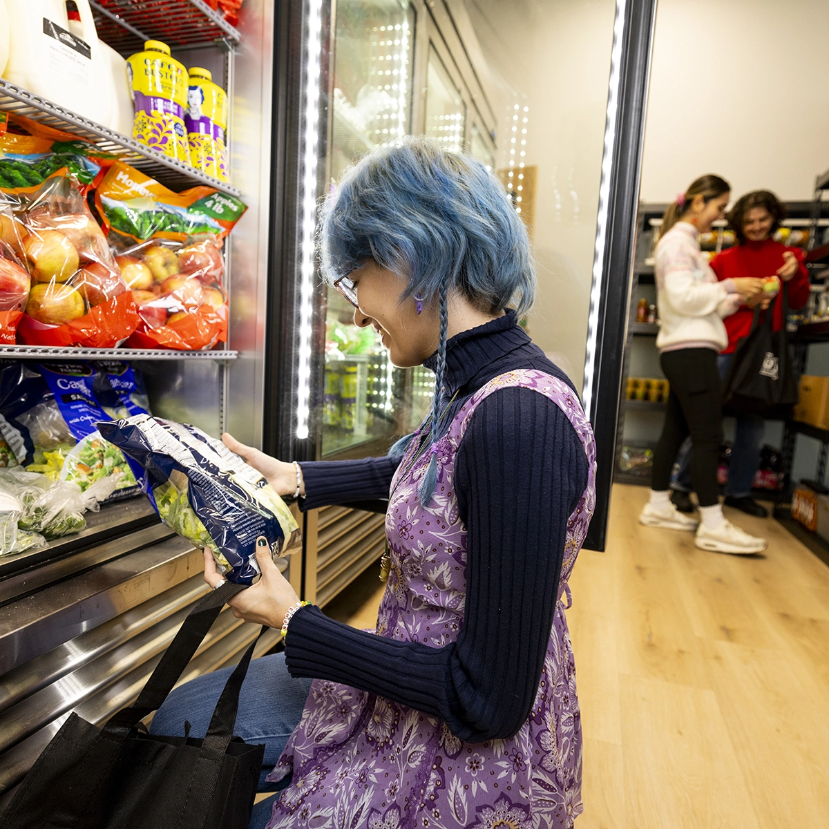 A woman with blue hair and wearing a purple dress reads the label on a bag of vegetables in the UVU CARE Hub’s student food pantry. A woman with blue hair and wearing a purple dress reads the label on a bag of vegetables in the UVU CARE Hub’s student food pantry.