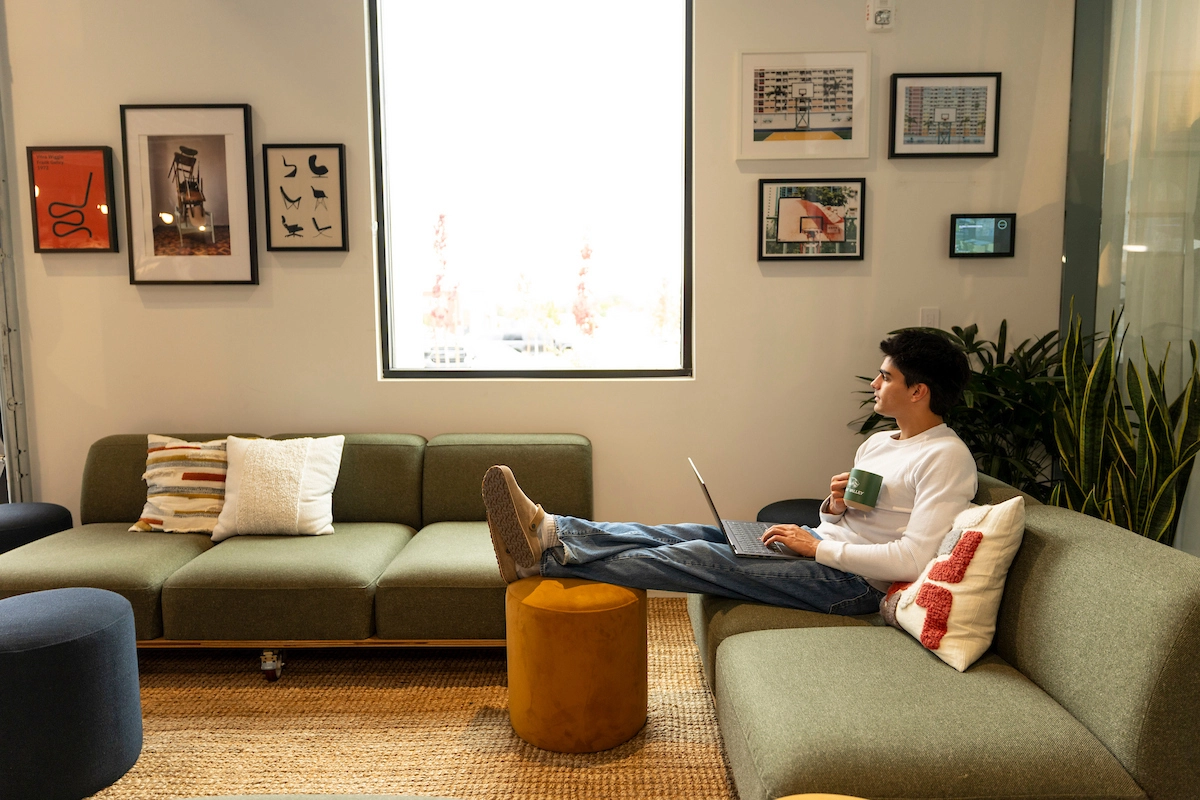 Student reclines on a green couch with a laptop. He has a drink in one hand and his feet up on an orange ottoman. Student reclines on a green couch with a laptop. He has a drink in one hand and his feet up on an orange ottoman.