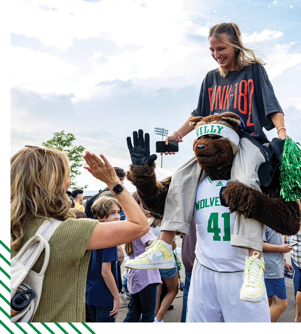 Willy the Wolverine high-fives a student in a green sweater as a student in a black shirt and khaki pants sits on his shoulders. Willy the Wolverine high-fives a student in a green sweater as a student in a black shirt and khaki pants sits on his shoulders.