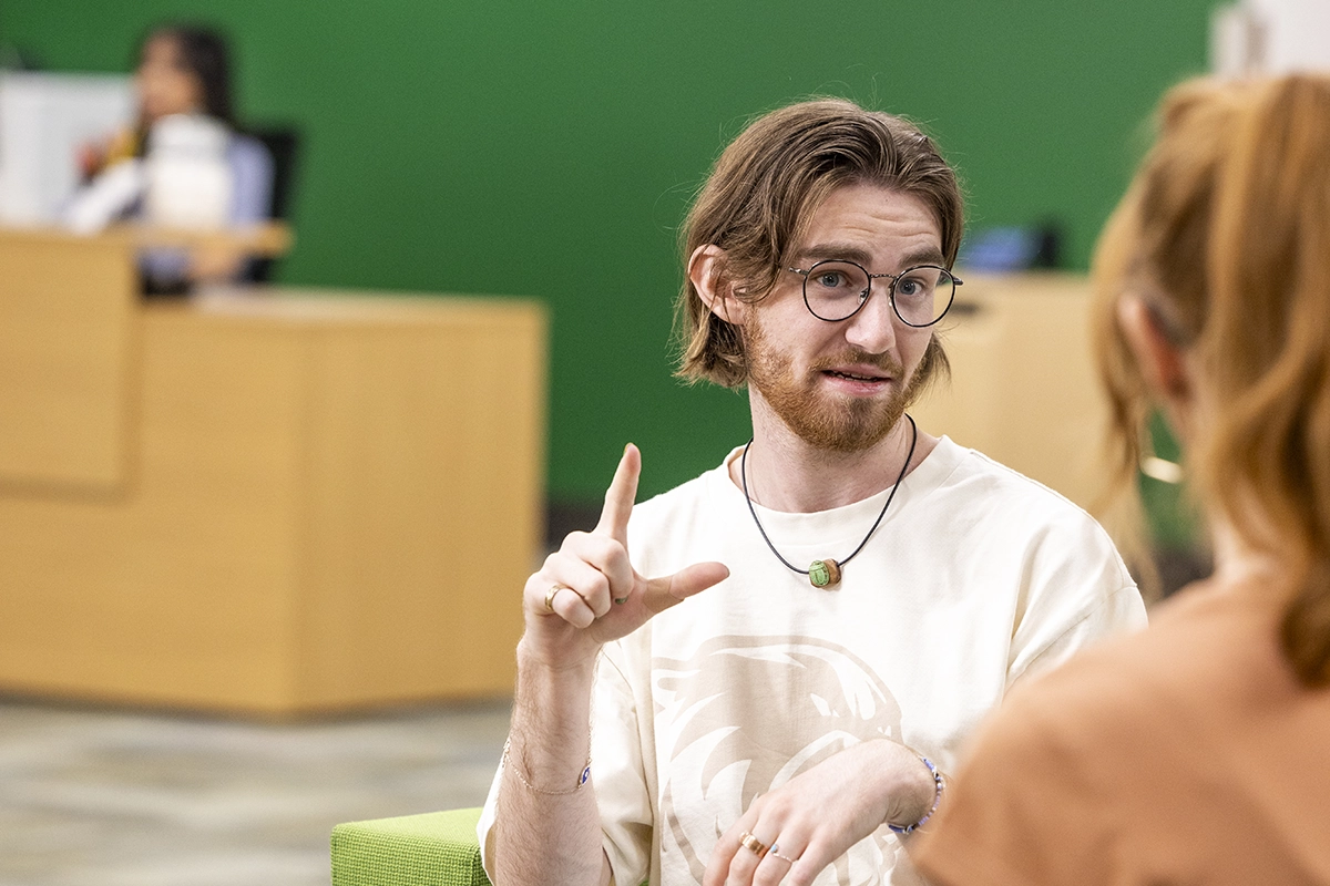A student with long brown hair and glasses sits in a green chair and communicates with another student using American Sign Language. A student with long brown hair and glasses sits in a green chair and communicates with another student using American Sign Language.
