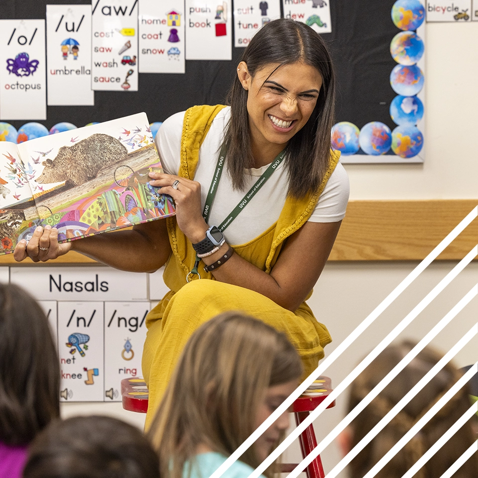 In an elementary school classroom, a UVU elementary education student reads a picture book to a group of children seated on the floor. In an elementary school classroom, a UVU elementary education student reads a picture book to a group of children seated on the floor.