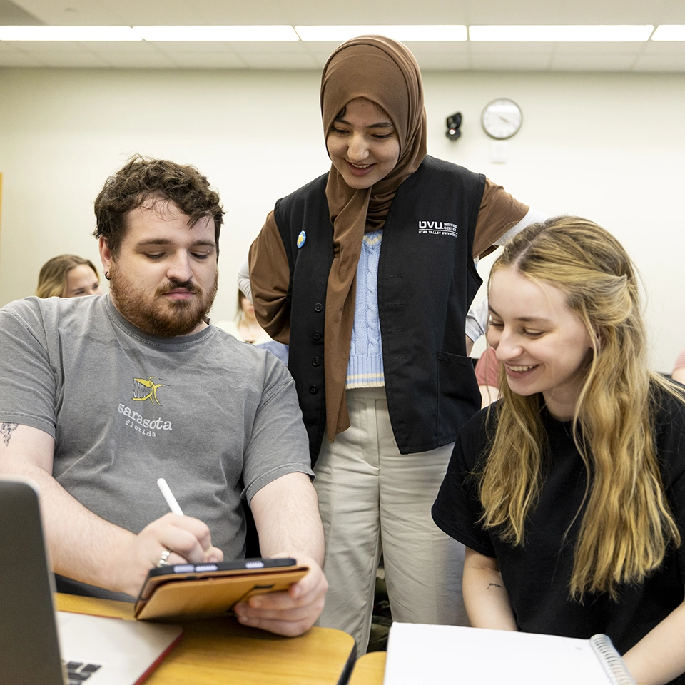 A UVU Writing Center instructor, wearing a black vest and a brown hijab, assists two students with their coursework. A UVU Writing Center instructor, wearing a black vest and a brown hijab, assists two students with their coursework.