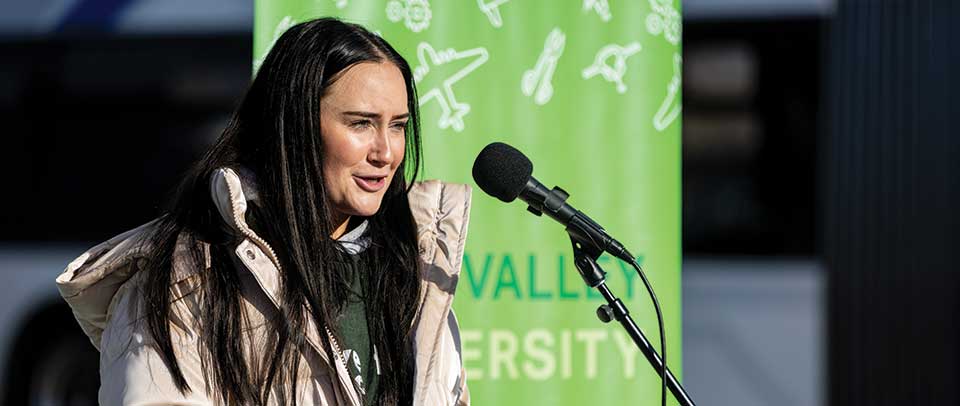 A UVU student, wearing a cream-colored coat and a green UVU T-shirt, speaks at a press conference at the Orem Central Station while announcing the “Give the Gift of Clean Air” campaign in partnership with UTA.