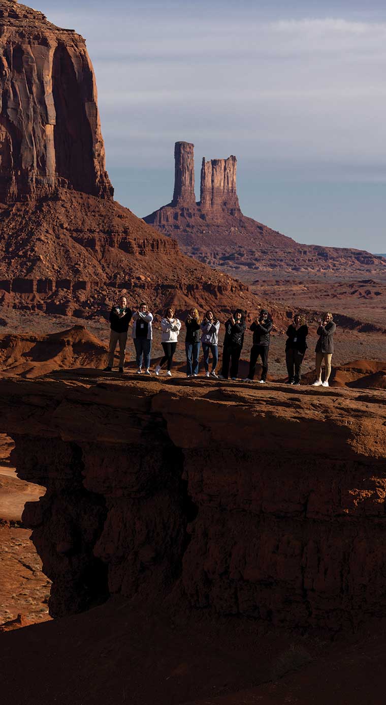 Nine students from UVU’s School of Education stand on a cliff in front of large rock formations in Monument Valley in Utah. 