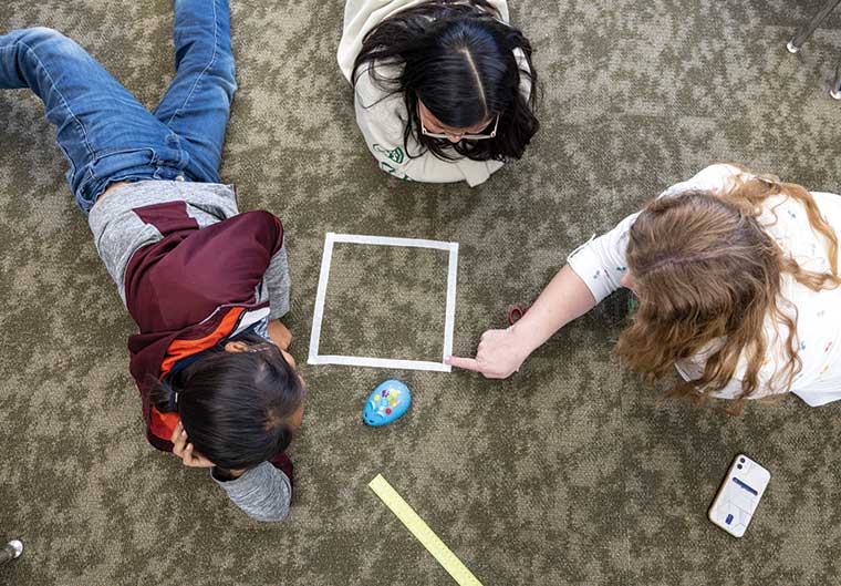A UVU School of Education student sits on the floor with two young girls from a Navajo Nation elementary school as they work with a robotic mouse. 