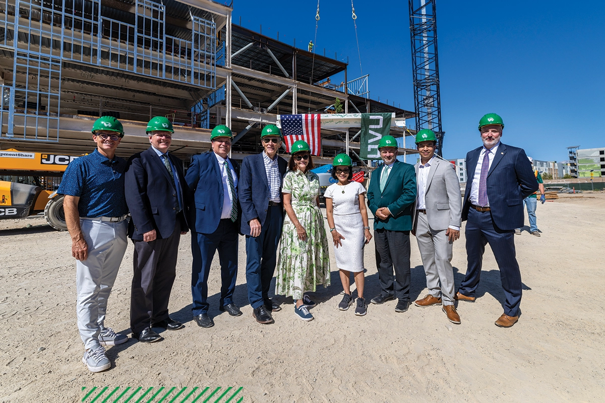 A group including President Tuminez, Scott and Karen Smith, Wayne Vaught, Kyle Reyes, Jim Mortensen, Val Peterson, and Kelly Flanagan wear green hard hats and pose in front of the Scott M. Smith Building, which is still being constructed. A group including President Tuminez, Scott and Karen Smith, Wayne Vaught, Kyle Reyes, Jim Mortensen, Val Peterson, and Kelly Flanagan wear green hard hats and pose in front of the Scott M. Smith Building, which is still being constructed.