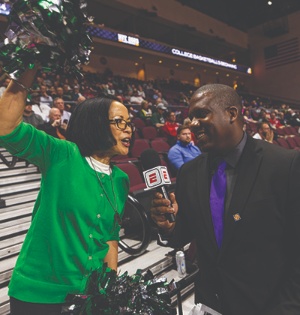 President Tuminez, wearing a green sweater, speaks with a reporter at an NIT basketball game. 
