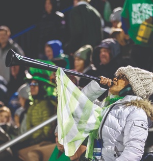 President Astrid S. Tuminez, wearing a winter coat and hat, blows a stadium horn at a Wolverine soccer game. 