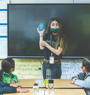 A UVU School of Education student, wearing a green face mask and a UVU T-shirt, stands in front of an elementary school class holding a robotic mouse, which is a teaching tool. 
