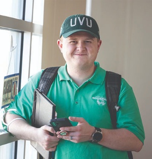 UVU staff member Nick Gledhill, wearing a green UVU polo shirt and a green UVU hat, smiles while at a Navajo Nation elementary school. 
