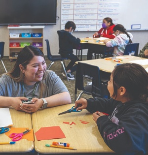 A UVU School of Education student smiles while working with students at a Navajo Nation elementary school, who are working with scissors, glue sticks, and construction paper. 