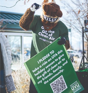 Willy the Wolverine, wearing a green T-shirt that says “Give the Gift of Clean Air,” holds a green poster bearing a pledge to promote sustainability by taking public transit