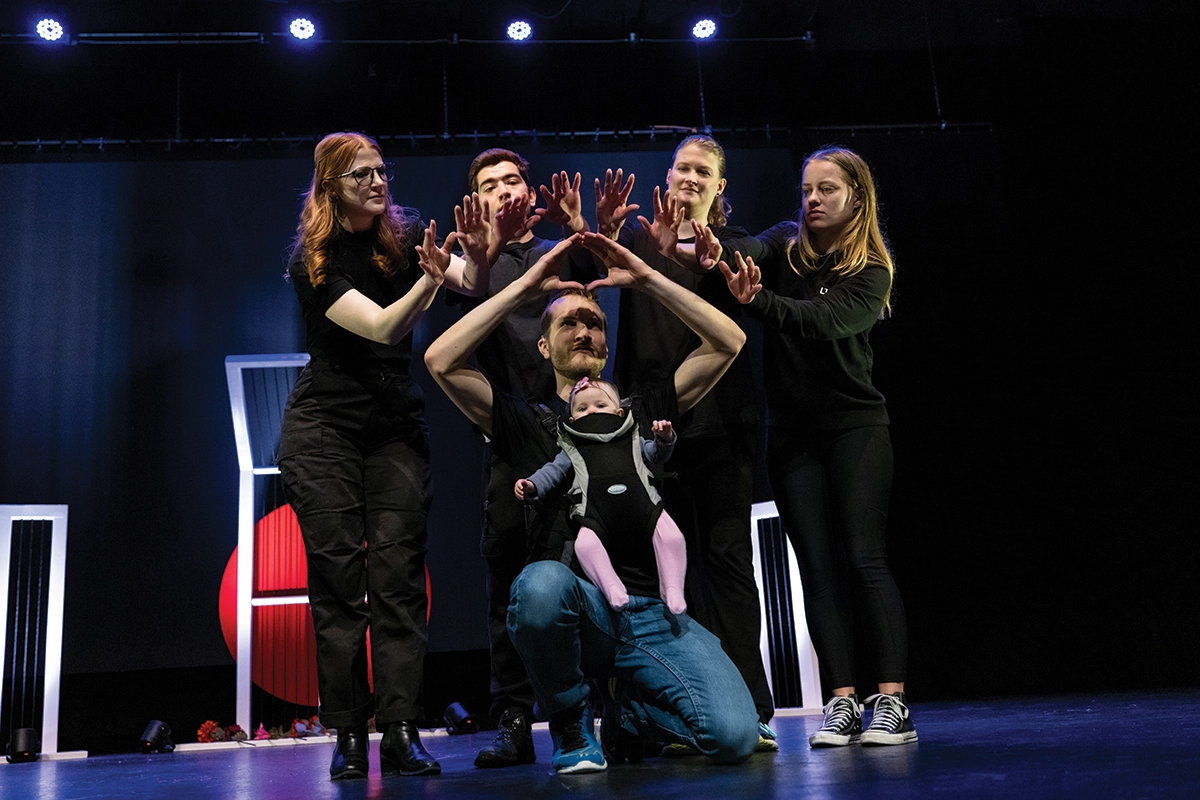 Five adults, one with a baby, sign American Sign Language on a stage