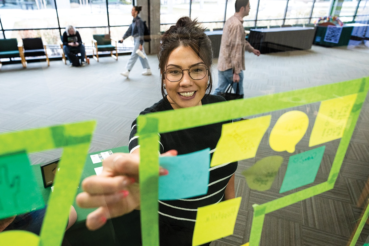 A student wearing glasses and a black and white striped shirt places a blue sticky note on a word wall celebrating native languages. 