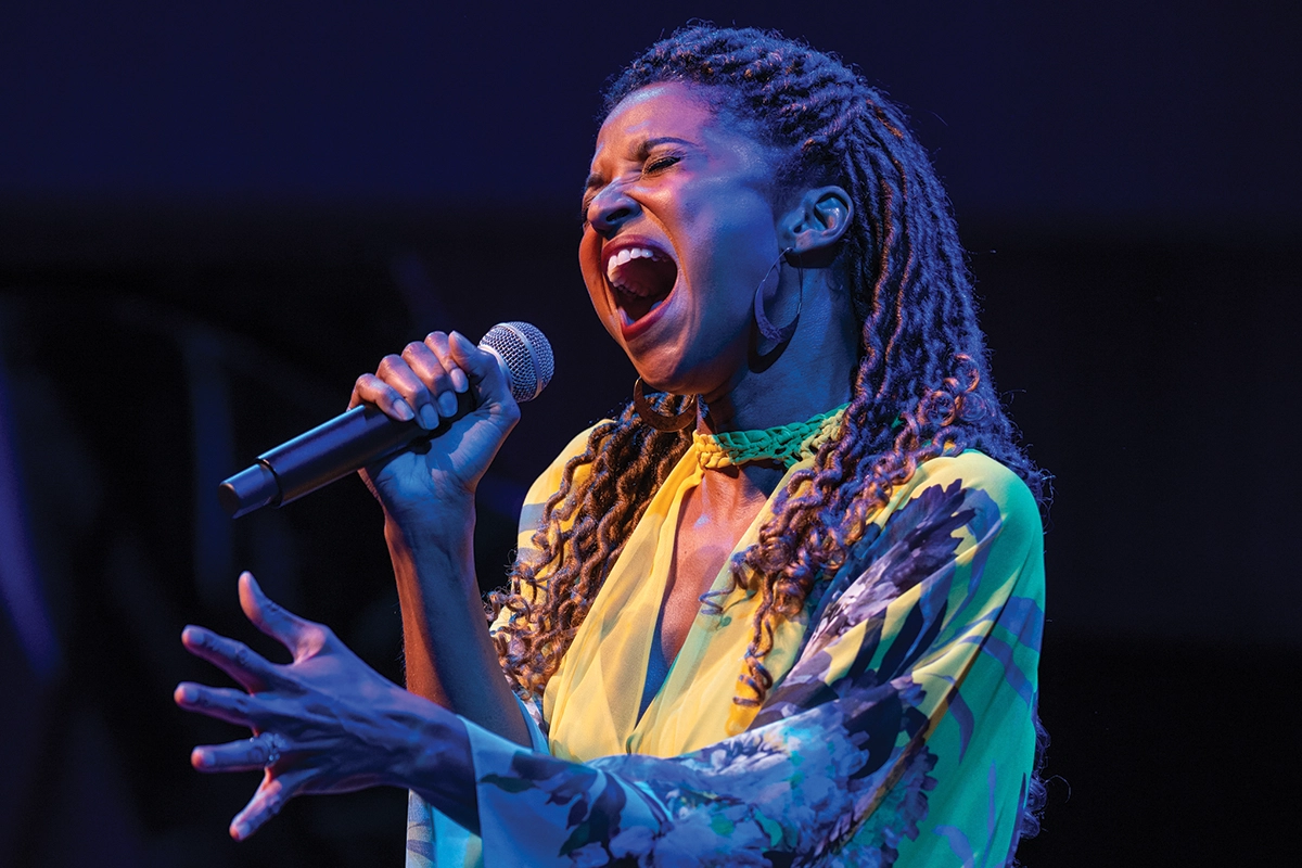 Broadway star Renée Elise Goldsberry, wearing a floral dress, sings into a microphone during an event at The Noorda Center for the Performing Arts. 
