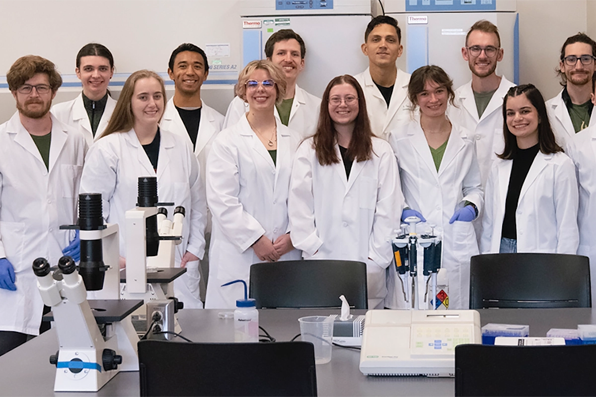 A group of UVU biotechnology students wearing white lab coats smile together in a UVU lab. 
