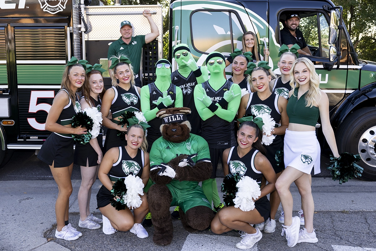 The UVU Green Man Group, the UVU cheer team, and the UVU dance team pose together in front of a UVU truck. 