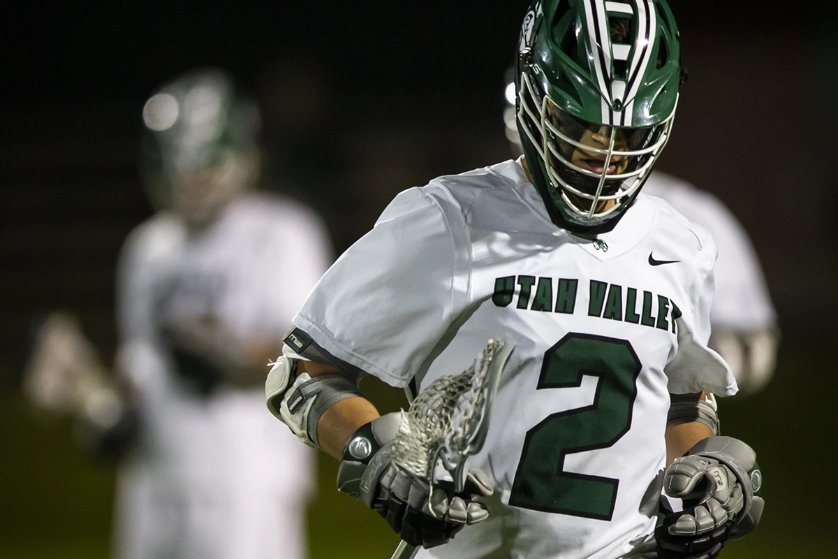 A Utah Valley lacrosse player, wearing a green and white jersey and a green helmet, plays on a lacrosse field. 