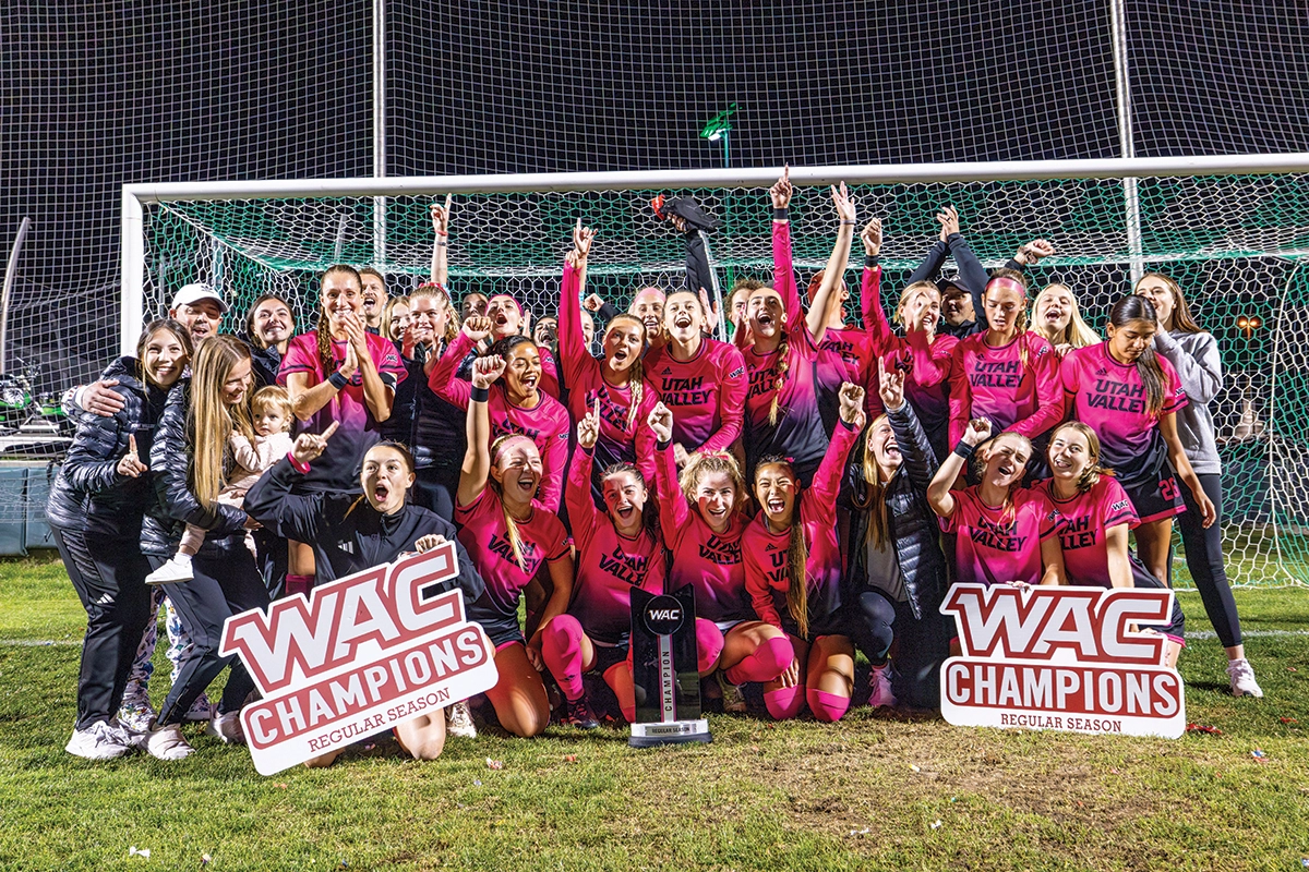The UVU women’s soccer team, along with coaches and family members, pose for a group photo with their WAC Championship trophy in front of a soccer goal on the field. 