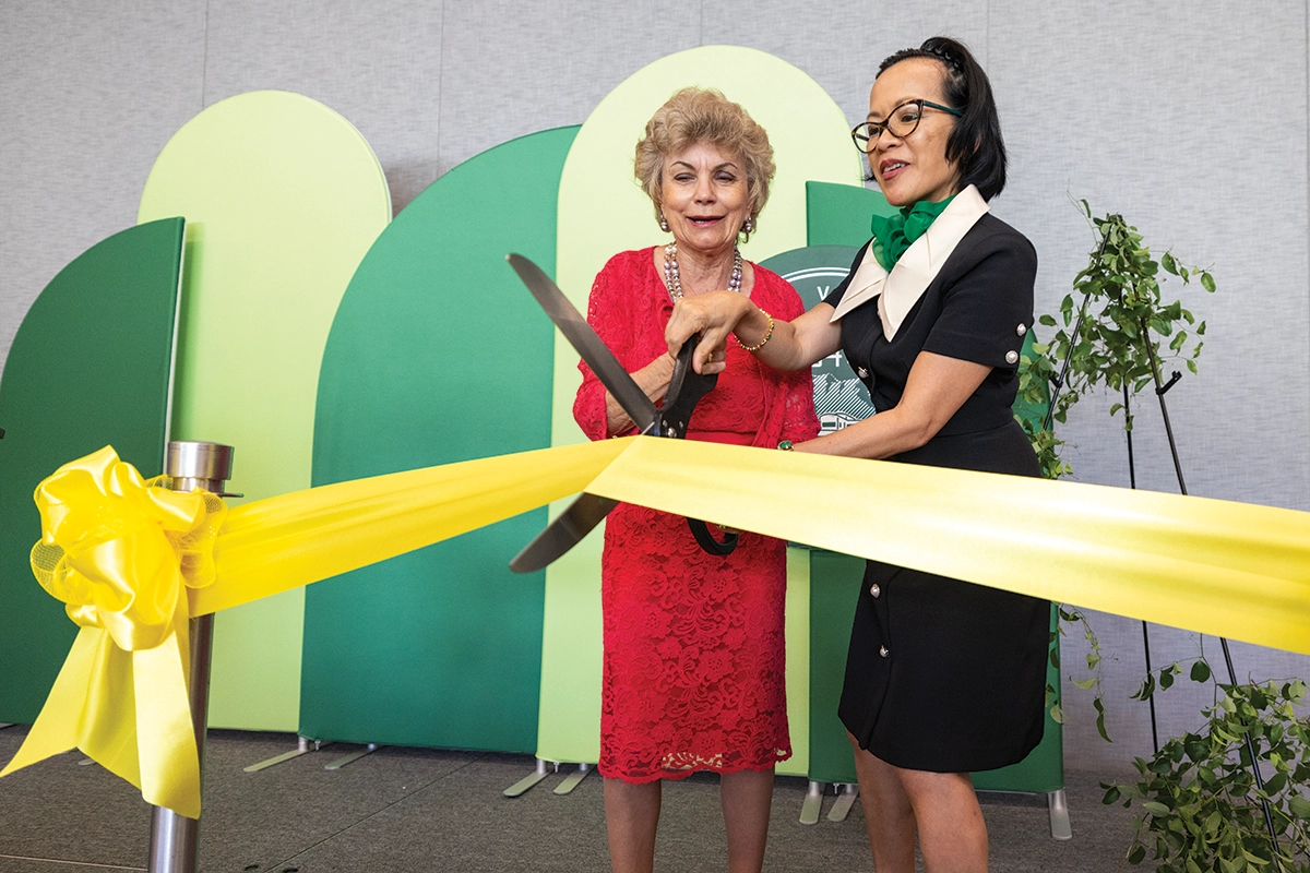 President Astrid S. Tuminez cuts a yellow ribbon with giant scissors alongside a woman in a red dress at the ribbon-cutting ceremony for the new Young Living Alumni Center building. 