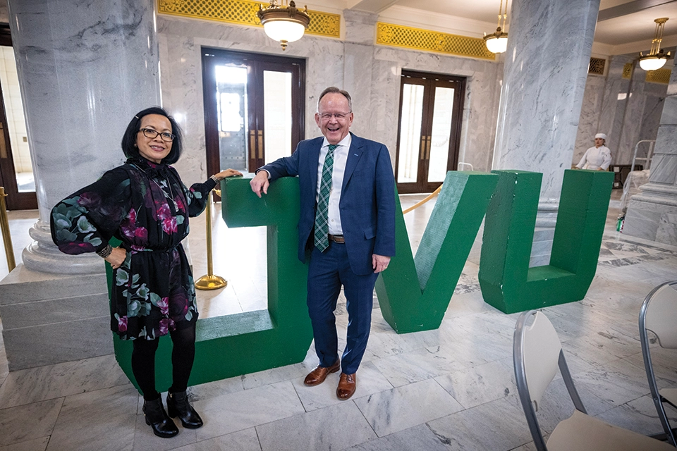 President Tuminez poses with a member of the Utah government in front of large, green UVU letters in the Utah Capitol Building. 