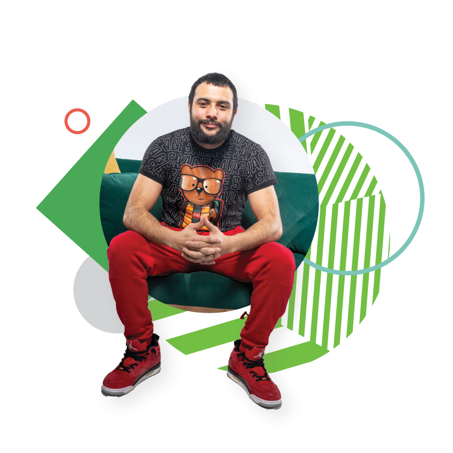 UVU student Pedro Del Valle, wearing a black graphic T-shirt, red pants, and red sneakers, poses while sitting on a green couch. 