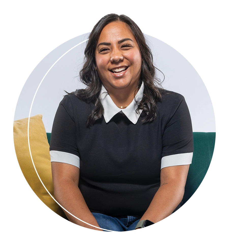 UVU alumna Kiana McAllister, wearing a black shirt with a white collar, smiles while sitting on a green sofa. 