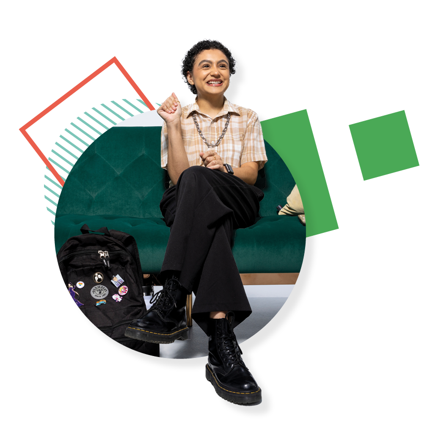 UVU alumna Priscilla VillaseÑor-Navarro, wearing a plaid shirt, black pants, and black shoes, smiles while sitting on a green couch.