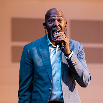 Thurl Bailey, wearing a gray suit, speaks into a microphone on stage. 