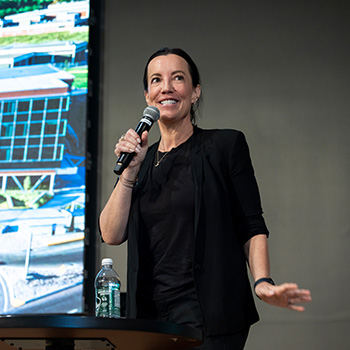 Jennifer Howell, wearing a black shirt and jacket, holds a microphone while lecturing on stage. 