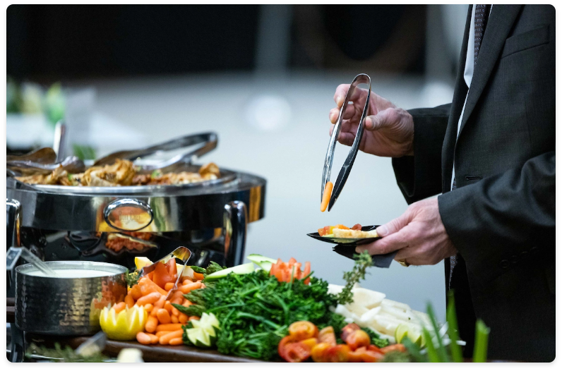 Person in a suit using tongs to pick vegetables from a buffet table with a chafing dish in the background.